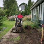 Mand kører minigraver og graver ud langs et hus i en have