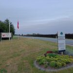 Indkørsel med skilt, blomsterbed og flagstang ved landevejen